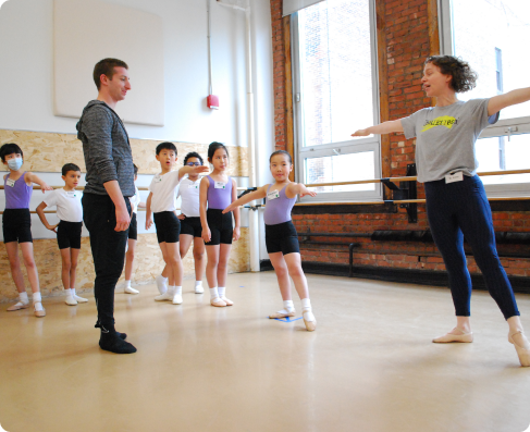 A group of young Introduction to Ballet students in a dance studio copy their teacher's movements, reaching one foot to a tendu side, and both arms outwards, preparing to dance across the floor.