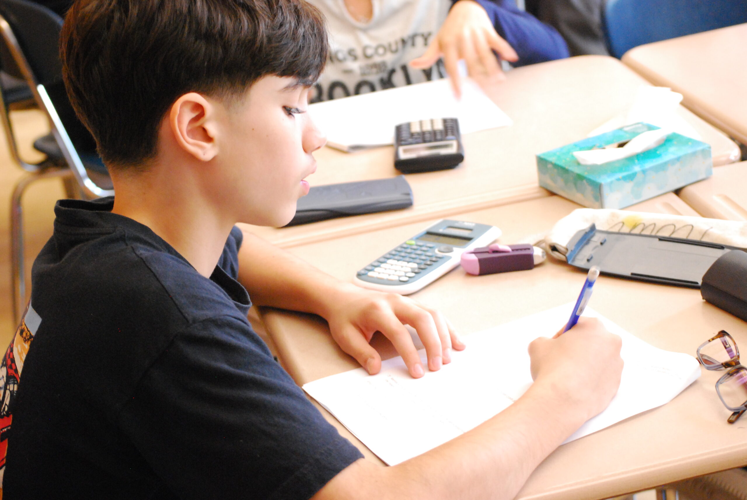 A student works at a classroom desk with a pencil, notebook, and calculator.