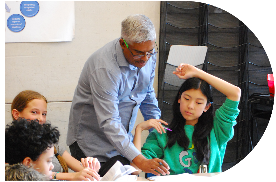 A teacher stands in a classroom hovering over a cluster of desks where students sit and work.