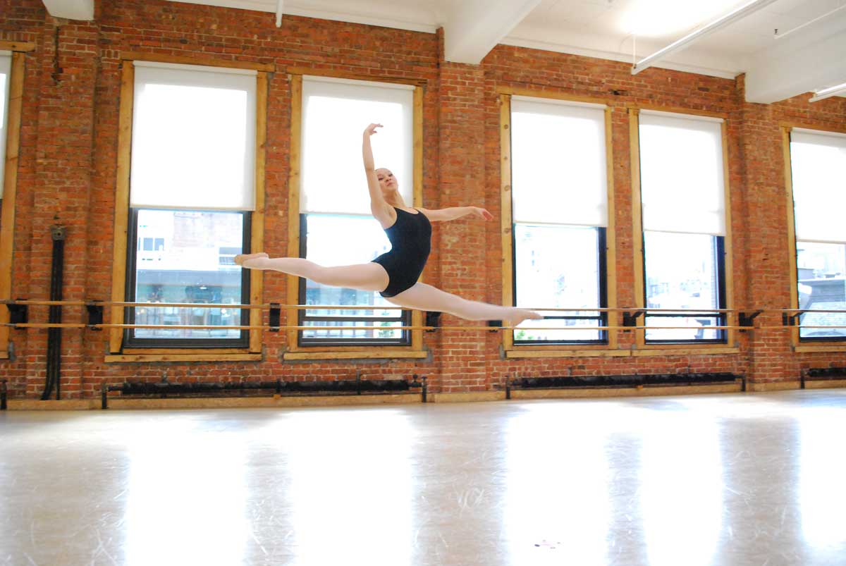 A teenage dancer in a ballet studio leaps across the floor in a split leap, with one arm framing her face and the other elongated to the side. Light streams through the windows.