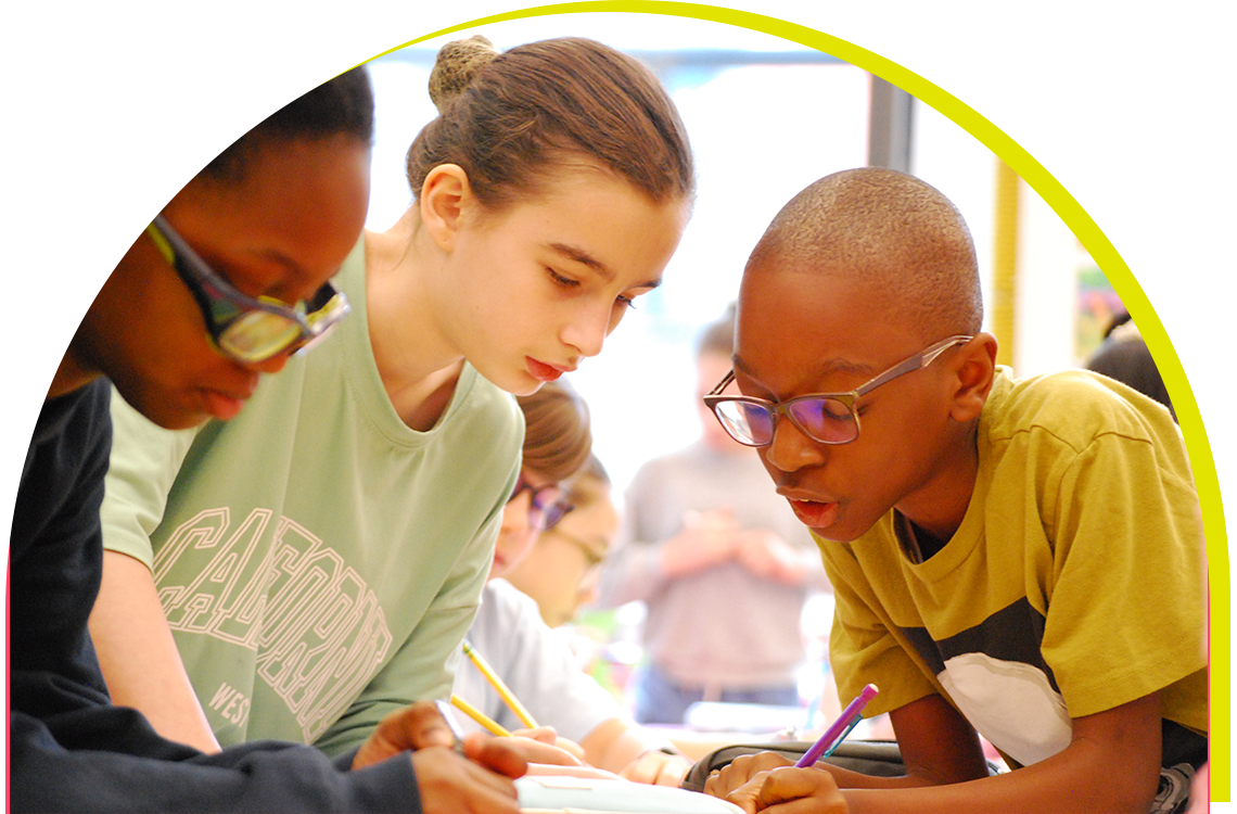 A group of three young students lean forward over their desks in a classroom, working on a project together.