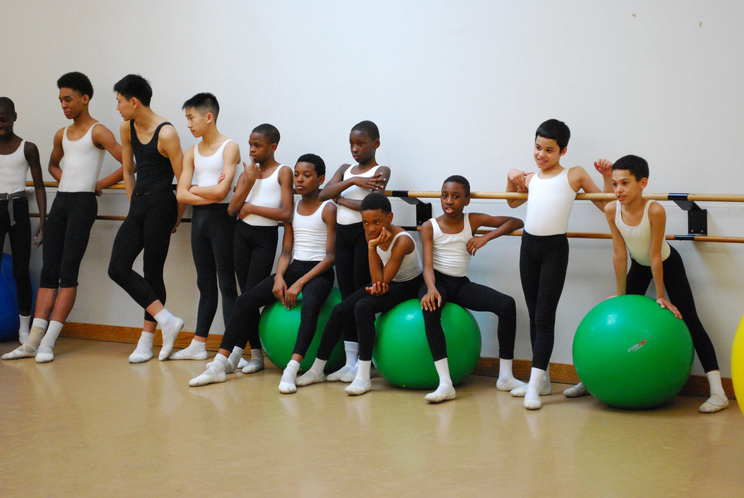 A group of young dancers in white leotards and black tights lean against the barre in the ballet studio in rehearsal. Some of them sit on green Pilates balls.