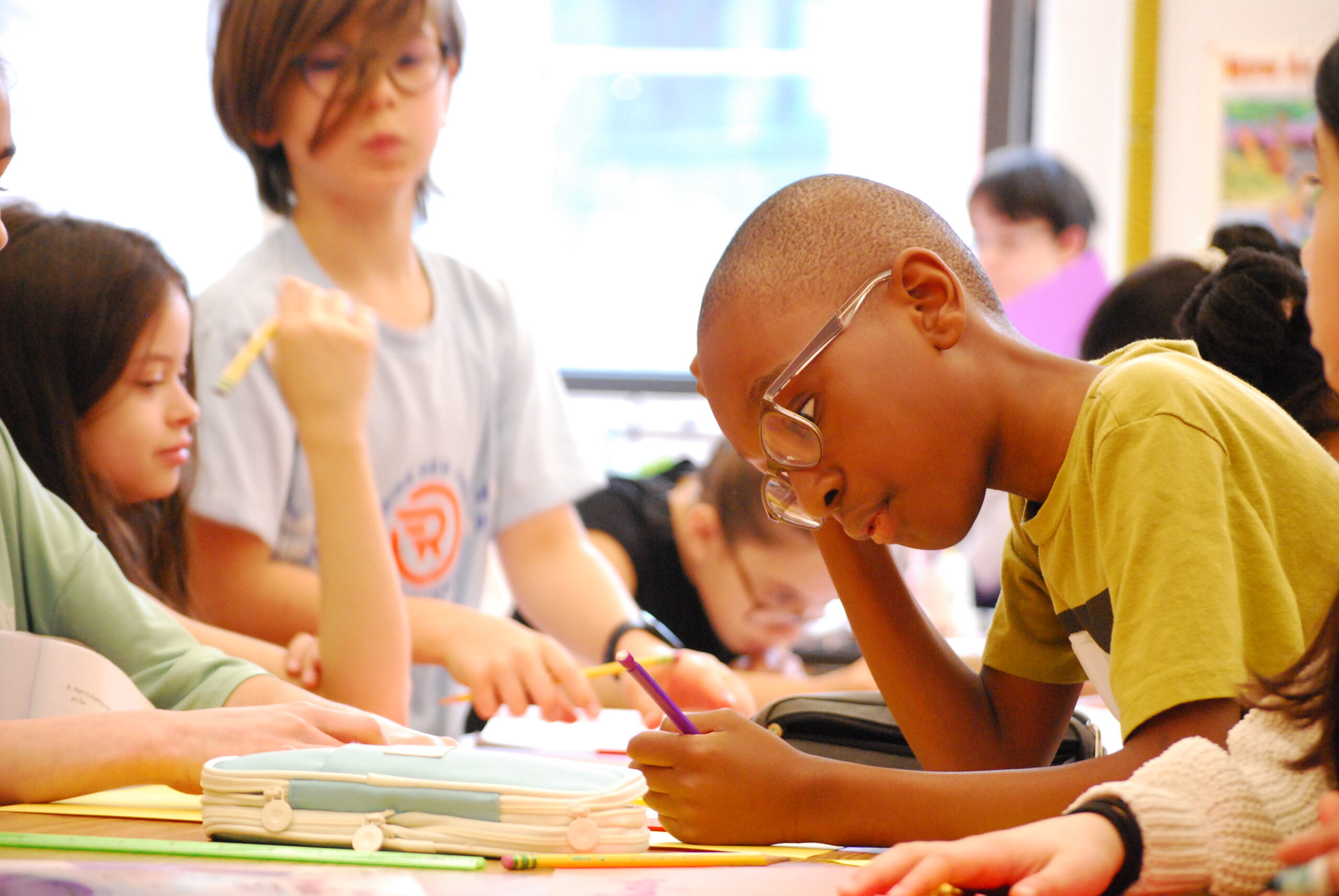 A student in a classroom wearing glasses rests his head on his hand as he works at his desk. Students work and smile in the background.