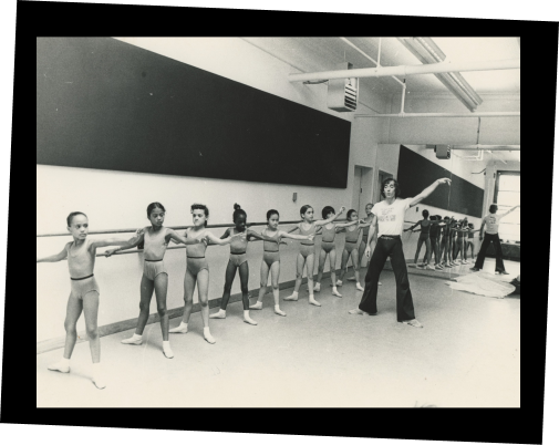 In a black and white photo, Eliot Feld teaches ballet class to a group of young students. They are doing plies in second position at the barre.