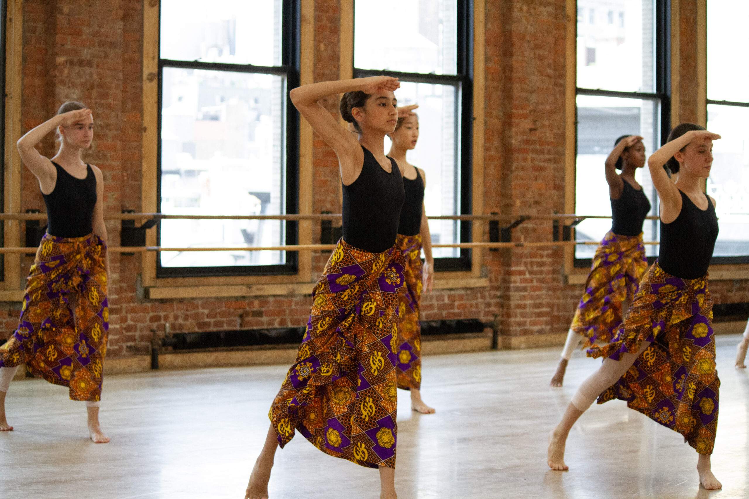 A group of students in a dance studio wear leotards with West African lapa skirts. They lunge forward on one leg, and touch one hand to their foreheads.