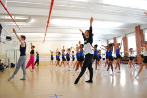 A group of young children in blue and white leotards copy their teachers at the front of the studio as they demonstrate a tendu forward from first position with arms in a v-shape. The teachers are from Parsons Dance Company, and are warming up the Elementary School students in rehearsal for Playscape Performs.