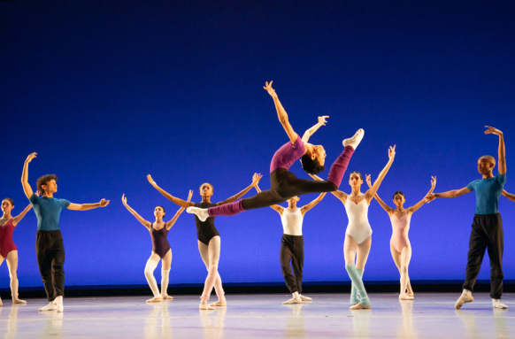 A lively ensemble of dancers dressed in bright, colorful costumes on stage, featuring a dancer in the center in a split leap with his head arched back.