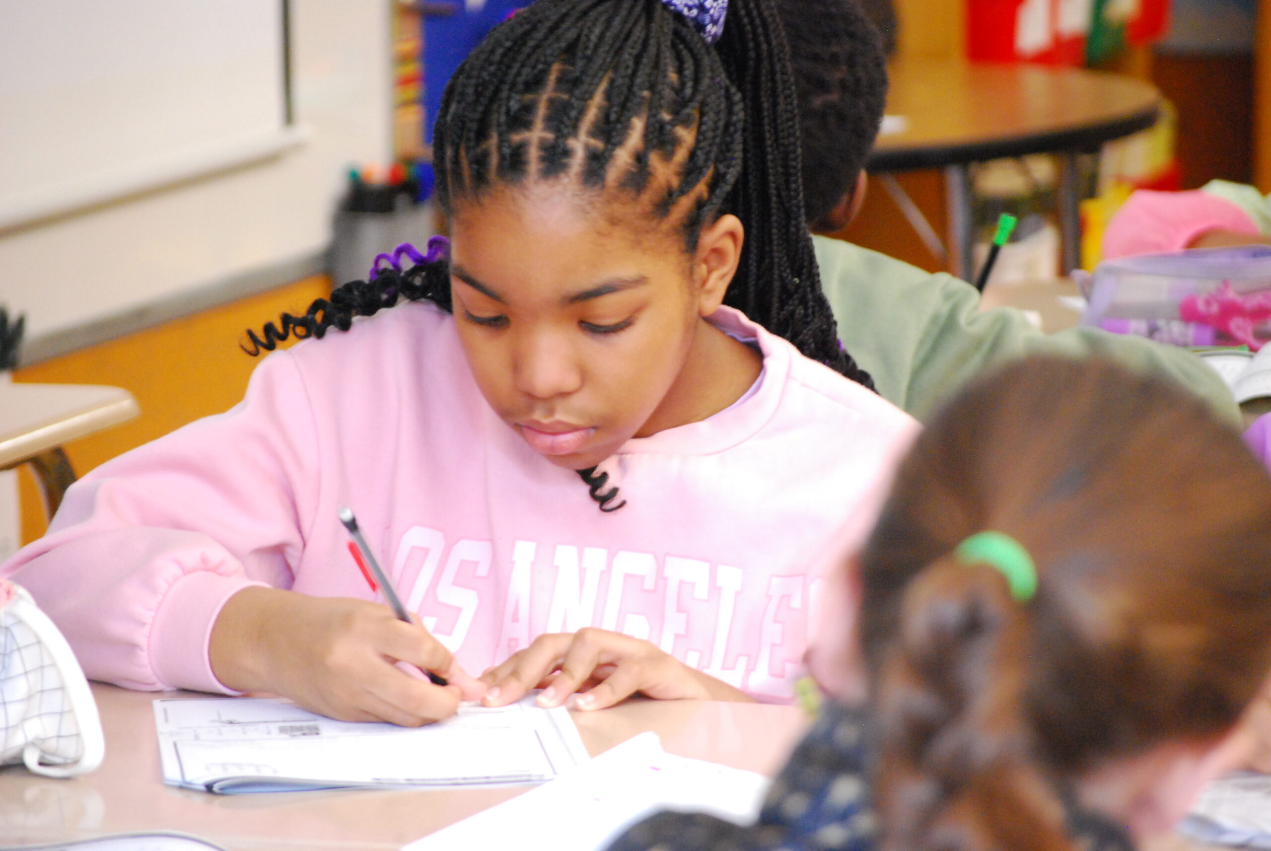 A young student in a classroom works at her desk, writing with a pencil.