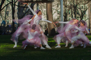 A group of dancers in white costumes with pink veils dance outside in a park, clustered together and reaching their arms outward, forming elegant and graceful shapes.