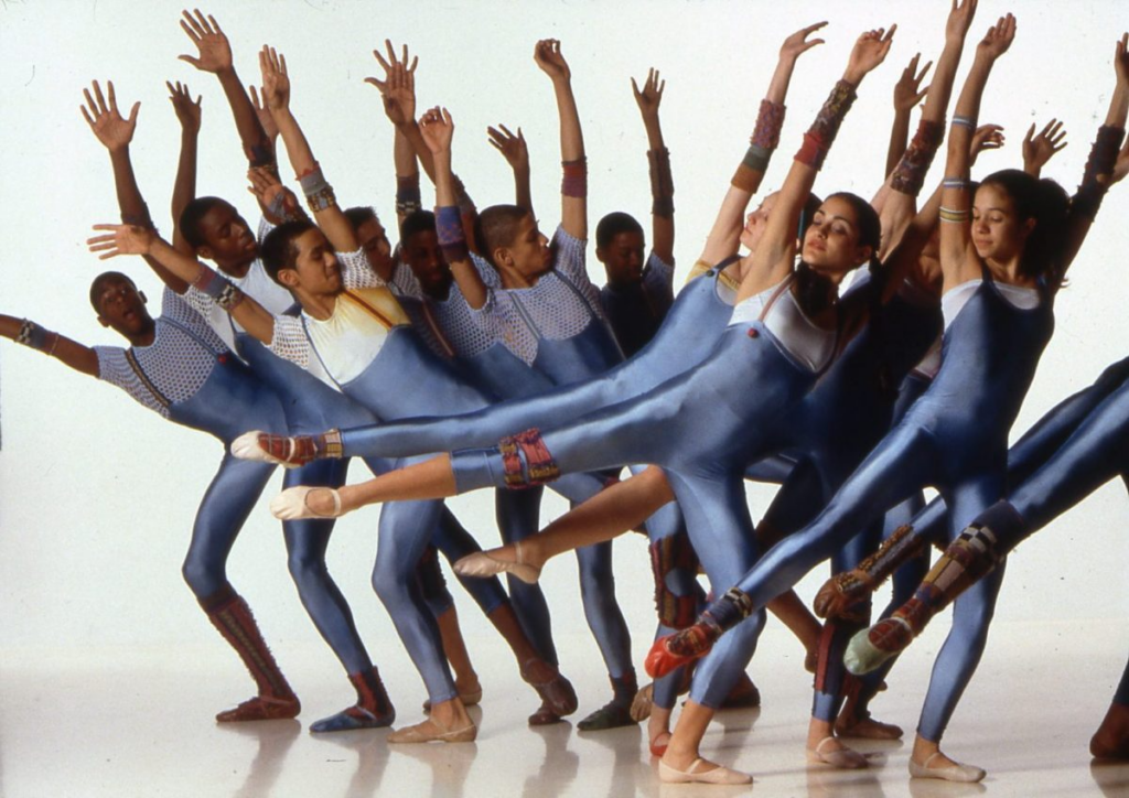 An ensemble of performers dance in two clumps, one in font of the other, wearing pale blue unitards and socks with funky patterns. They reach their arms above their heads in opposite directions, stretching horizontally from their fingers to their toe, extending a leg to the side.