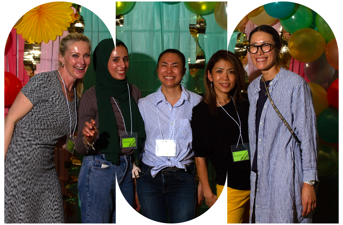 Five parents stand together smiling in front of a colorful backdrop with balloons. They wear visitors passes, participating at a school event.