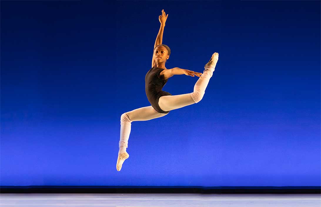 A ballet dancer in a black leotard and white leg warmers jumps gracefully with legs in a stag split and arms extended. The background is a gradient of blue shades, emphasizing her mid-air pose and expression of concentration.
