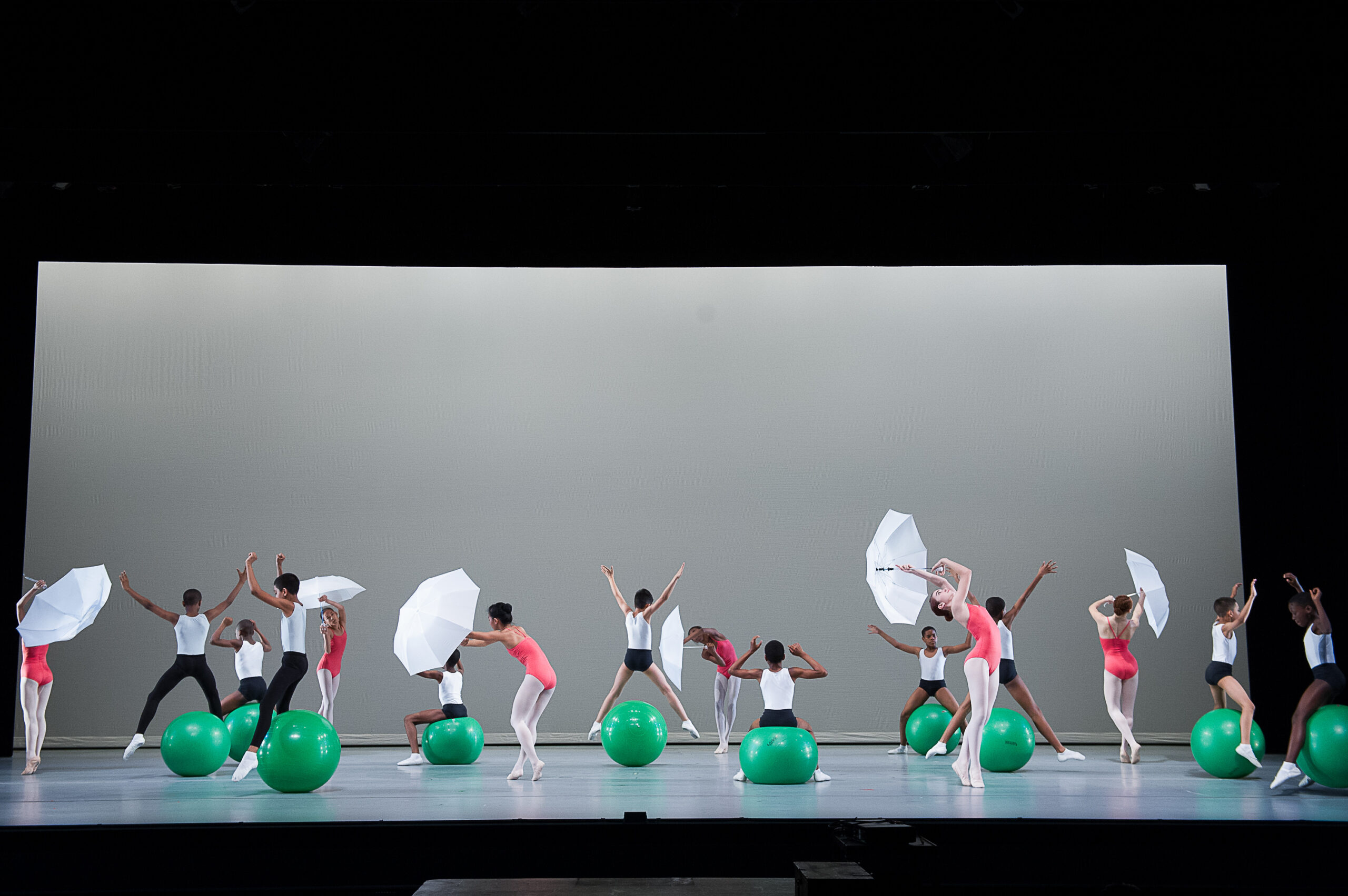 Dancers perform on stage with large green exercise balls and white umbrellas. They are arranged in a broad formation, some standing and others seated on the balls, all mid-movement. The background is a minimalist white while the stage is illuminated brightly.