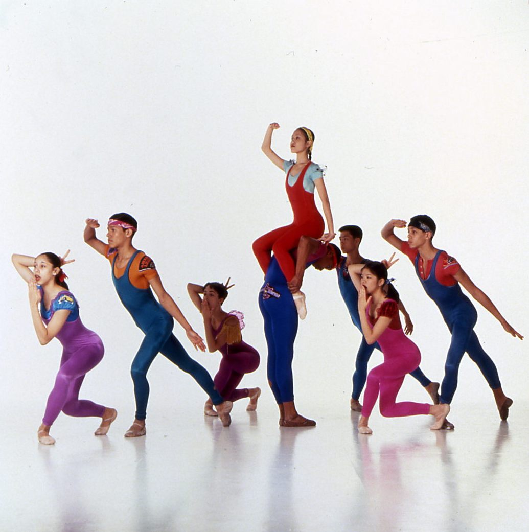 A group of eight dancers in colorful, form-fitting outfits perform a dynamic dance routine. One dancer is elevated on another's shoulders at the center. The dancers strike various expressive , forward leaning poses against a plain white background.