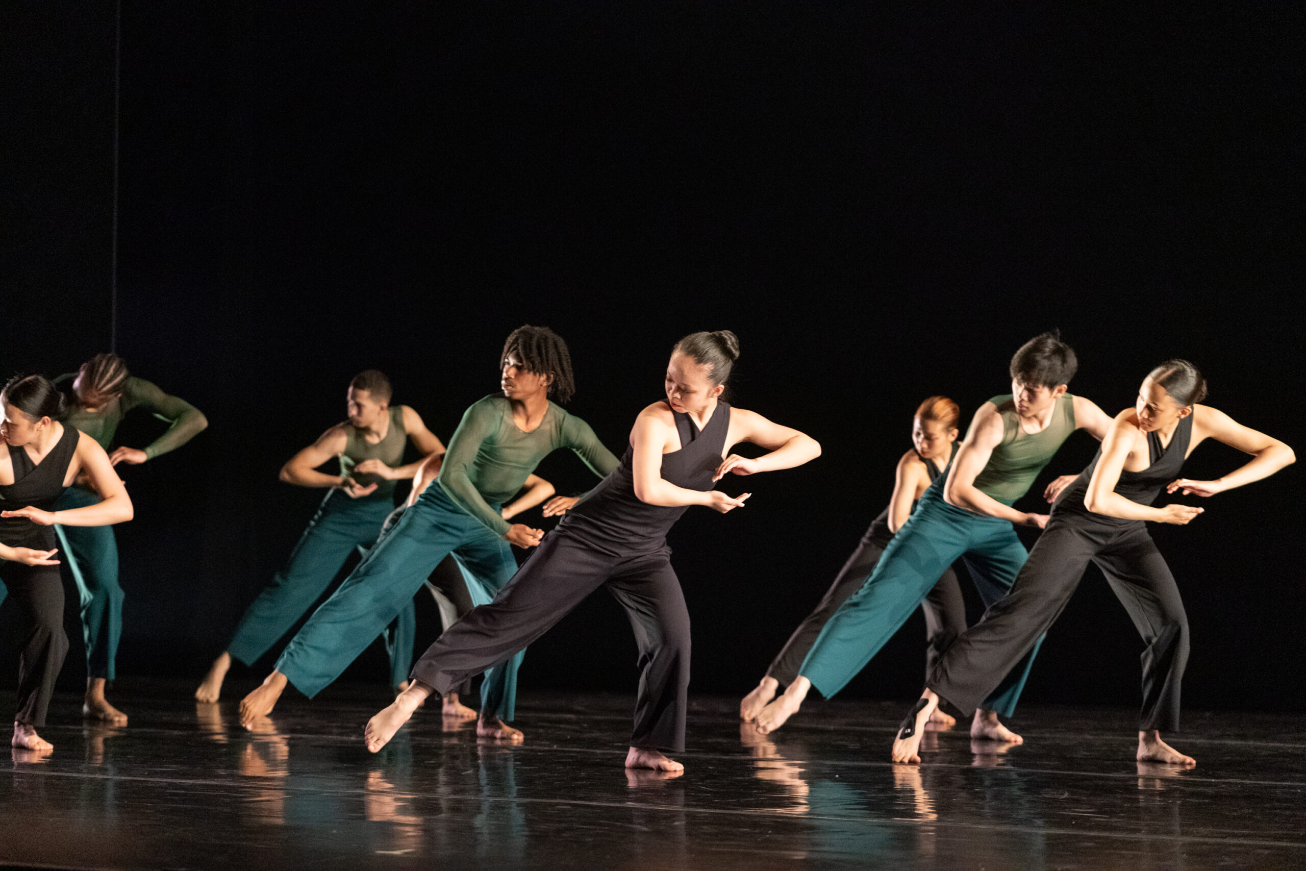 A group of dancers performs on stage, each wearing green and black costumes. They move in unison, holding their hands cupped atop one another with one leg extended to the side against a dark background.
