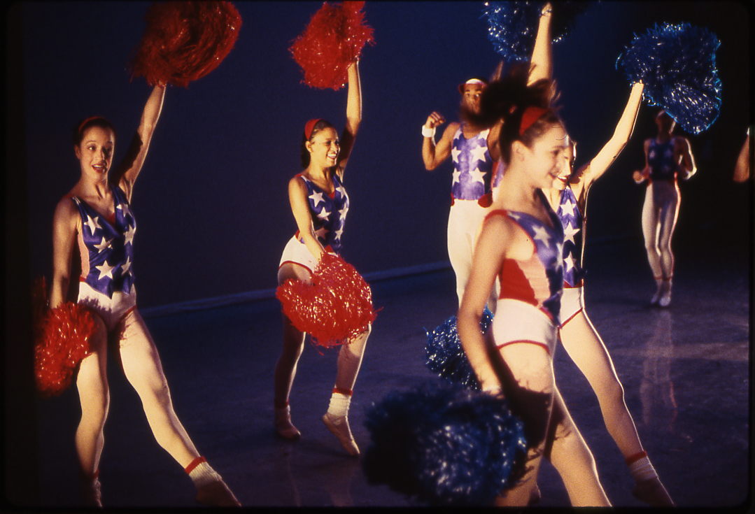 A group of dancers dressed as cheerleaders perform a routine. They are wearing patriotic outfits with American flag patterns and holding red, white, and blue pom-poms. The background is dark, highlighting the bright colors of their costumes and accessories.