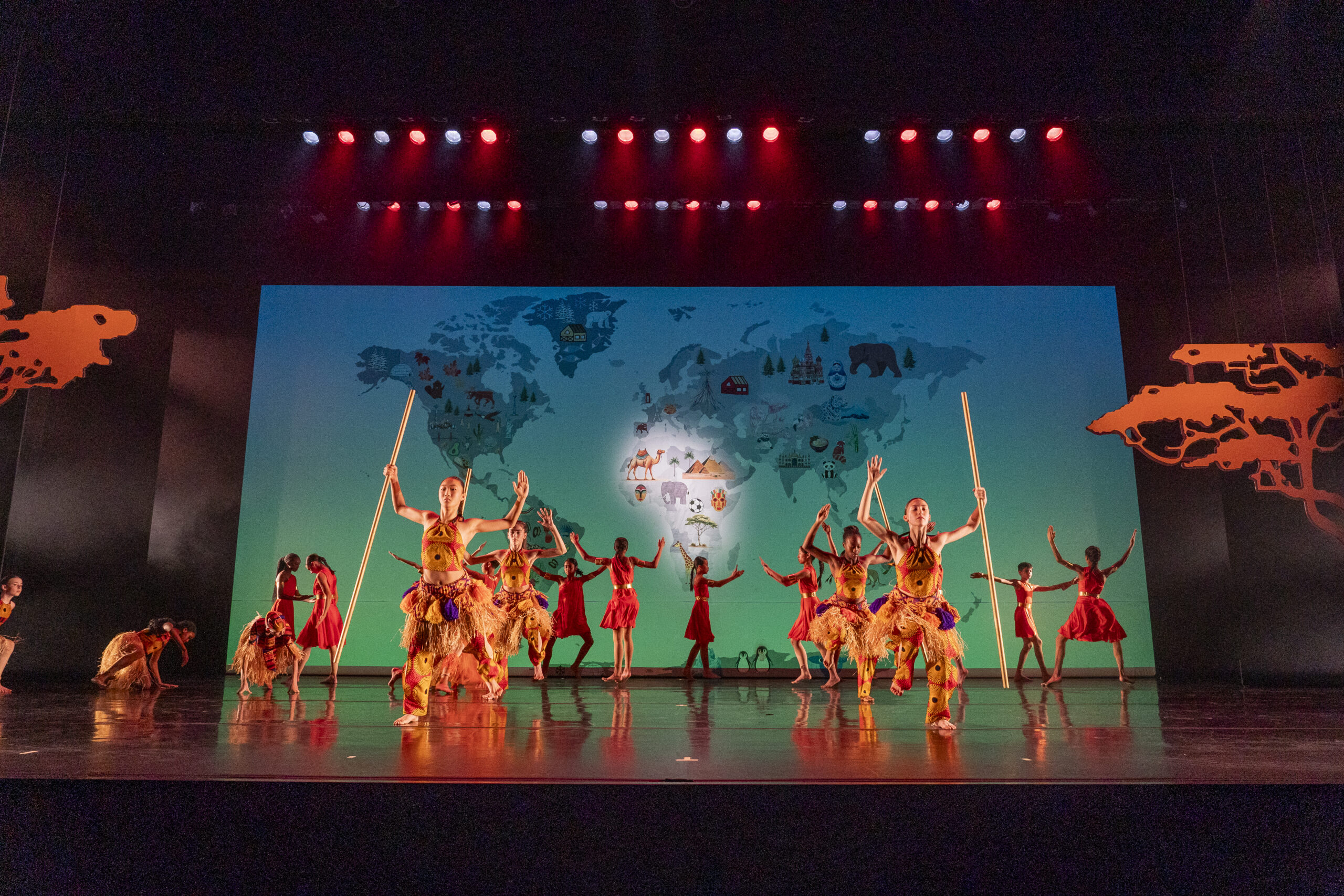 A group of dancers in colorful West African costumes performs on a stage with a backdrop displaying a world map. The stage is lit with red and blue lighting, and dancers hold long staffs while striking dynamic poses. Silhouettes of trees are on each side of the stage.