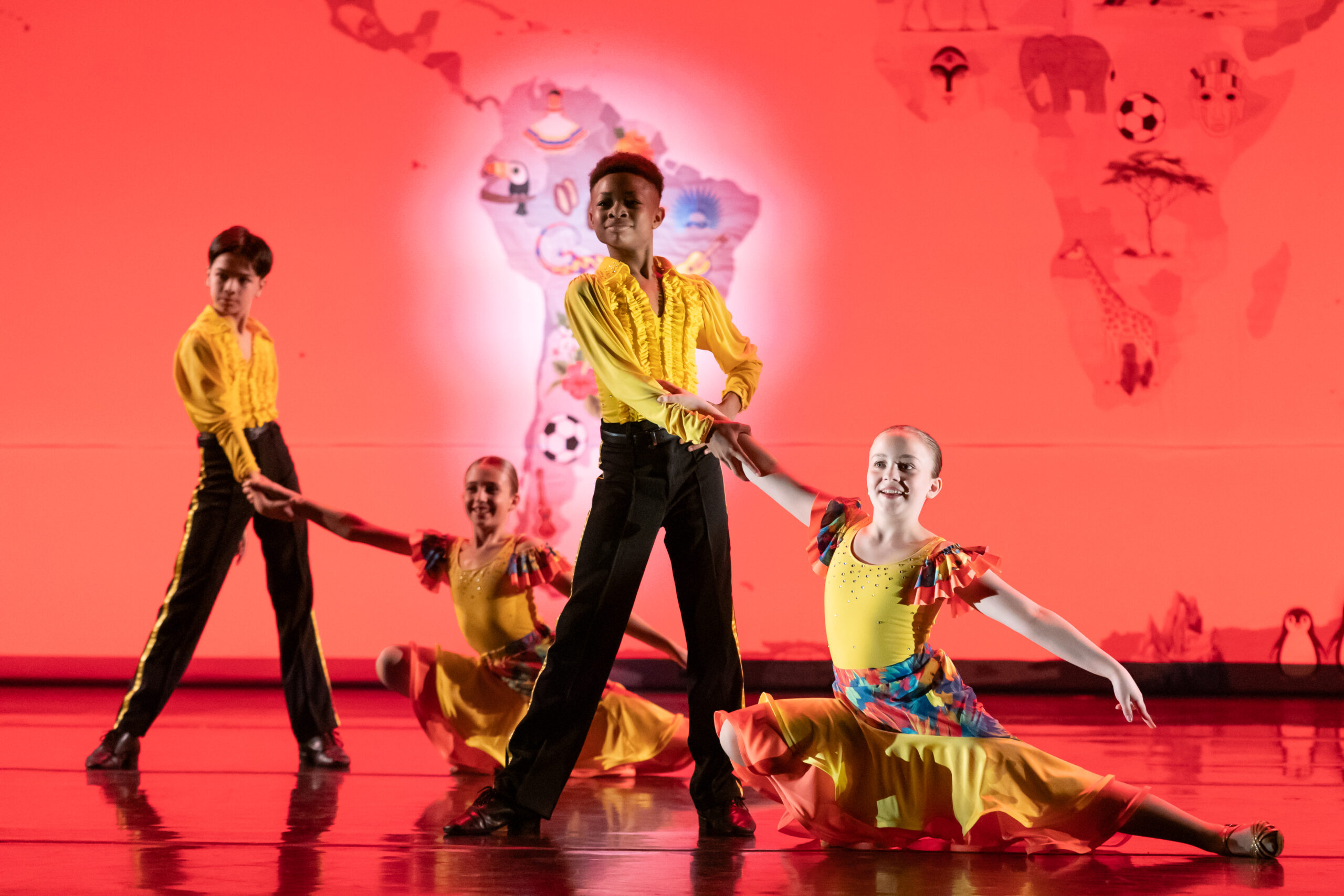 Four dancers in yellow tops and vibrant skirts and pants perform a lively dance on stage. The background features a red, illuminated map of the world with notable icons and landmarks. Two dancing couples showcase a mid-pose hold, with smiles highlighting their enthusiasm.