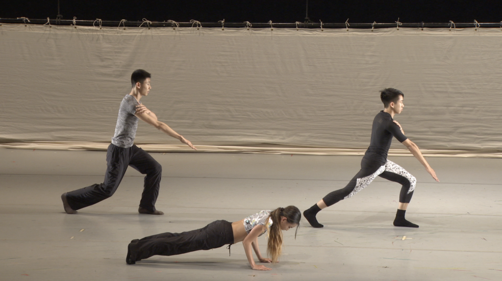 Three dancers dressed in neutral tones are seen on stage performing contemporary movements. The two upstage dancers are in a lunging position with arms extended, while the downstage dancer is in a plank position.