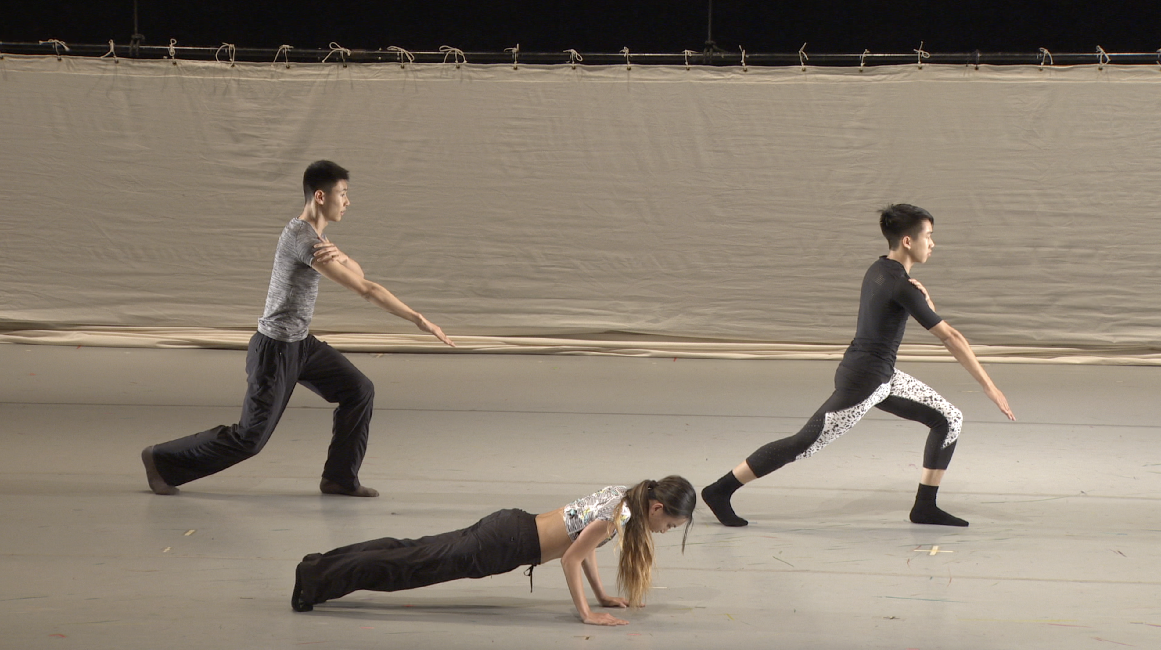 Three dancers dressed in neutral tones are seen on stage performing contemporary movements. The two upstage dancers are in a lunging position with arms extended, while the downstage dancer is in a plank position.