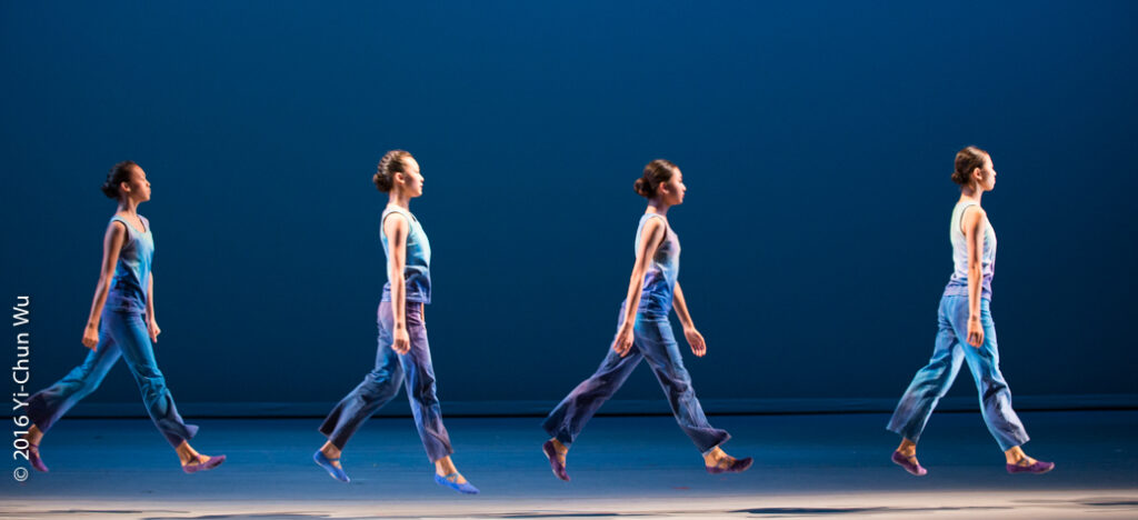 Four dancers in matching blue outfits jump in unison on a stage with a blue background. Each dancer extends one leg forward as they appear to be jumping in mid air with synchronized steps. The floor is smooth and the lighting is bright.