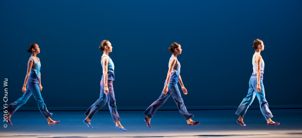 Four dancers in matching blue outfits jump in unison on a stage with a blue background. Each dancer extends one leg forward as they appear to be jumping in mid air with synchronized steps. The floor is smooth and the lighting is bright.