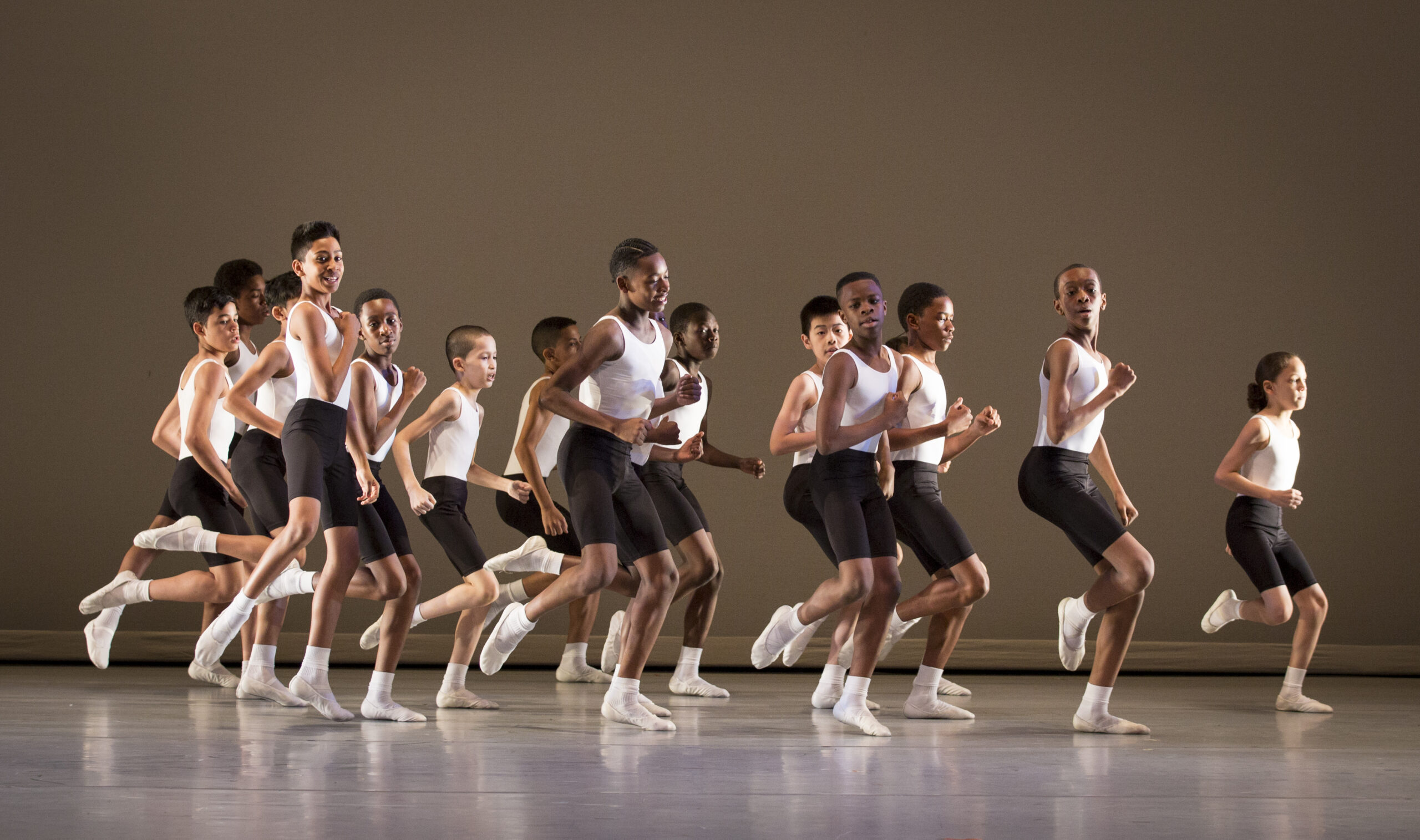 A group of students perform Eliot Feld's "Quickstep" on stage. Wearing white leotards with black shorts and white ballet slippers, a group of young boys jog in unison.