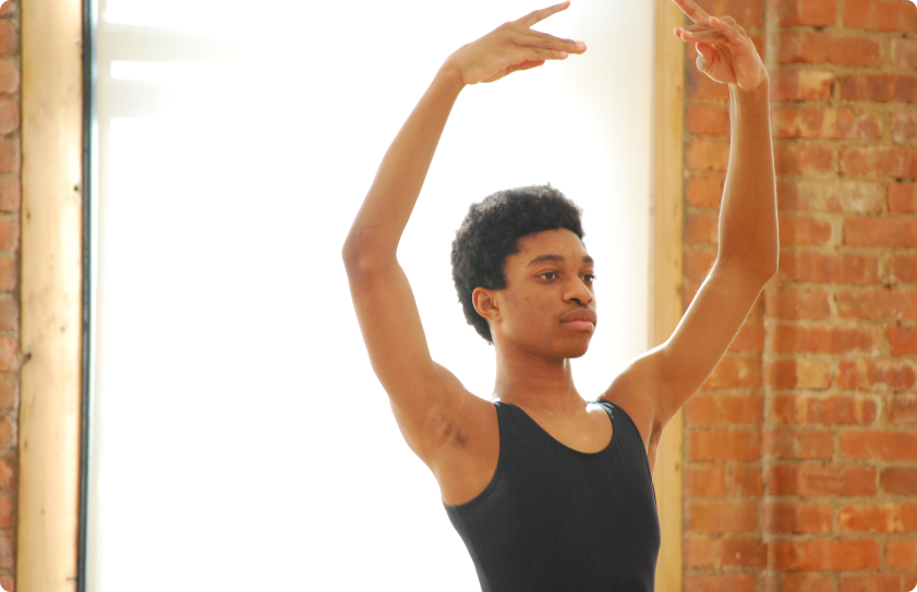 A boy in a ballet studio, shown from the waist up, looking focused as he raises his arms in 5th position.
