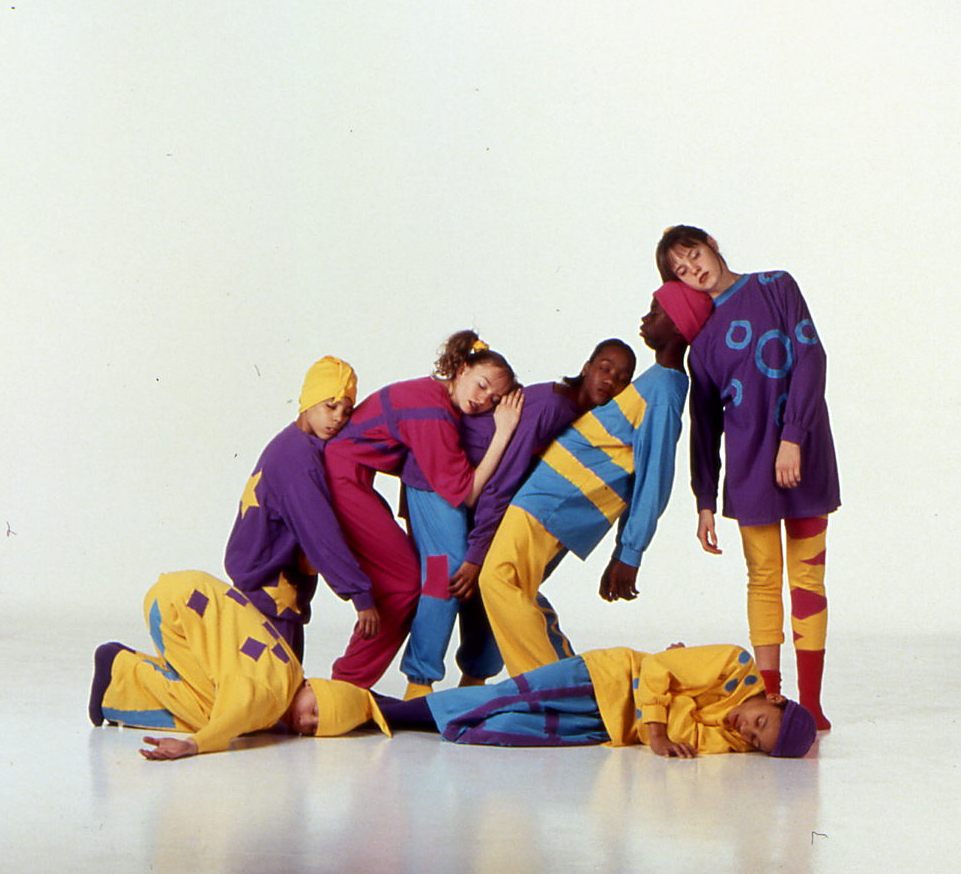 A group of six individuals pose in colorful, abstract outfits featuring bright yellow, blue, purple, and red patches. They stand and lie on a white background, positioning themselves in playful and relaxed sleeping poses that convey a sense of creativity and fun.