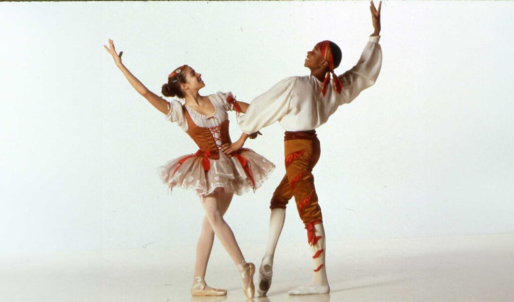 Two ballet dancers in costume strike a pose. The female dancer wears a white and red tutu with floral accents, while the male dancer is dressed in a white shirt, red pants, and a red headband. Both have one arm raised gracefully and the other interlocked.