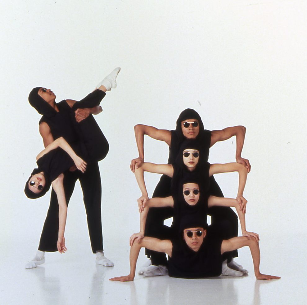 A group of four performers dressed in black form a pyramid-like structure with their bodies. Two performs stand next to the pyramid, one holding the other horizontally. All wear sunglasses and white socks.
