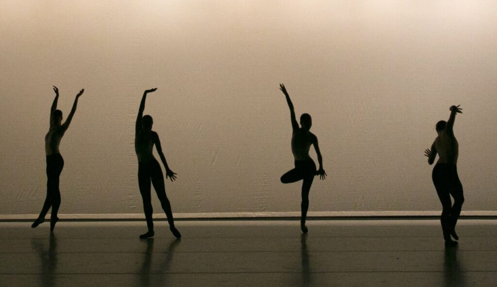 Four dancers wearing pointe shoes are in silhouette on a stage. They dance in a line, moving through graceful balletic gestures.