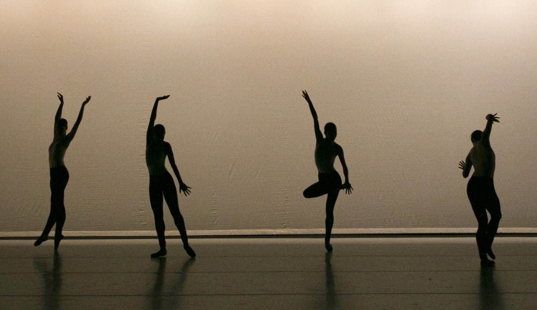 Four dancers wearing pointe shoes are in silhouette on a stage. They dance in a line, moving through graceful balletic gestures.