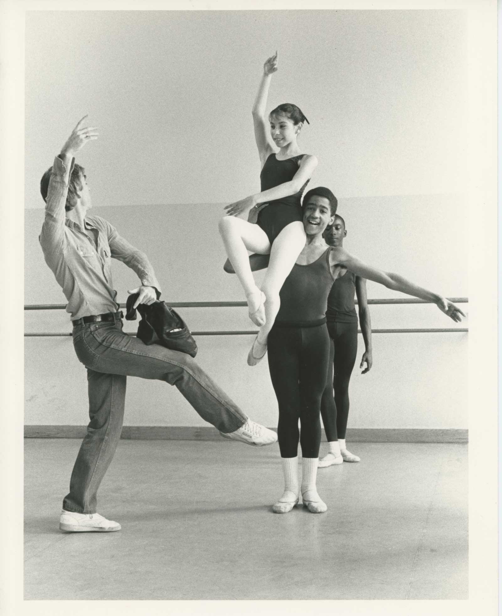 In a black and white photo, Eliot Feld demonstrates a front attitude pose in a dance studio, facing profile to the camera. His students look on; a boy lifts a girl onto his shoulder, and she mimics Mr. Feld's position from atop her perch, smiling down at him.