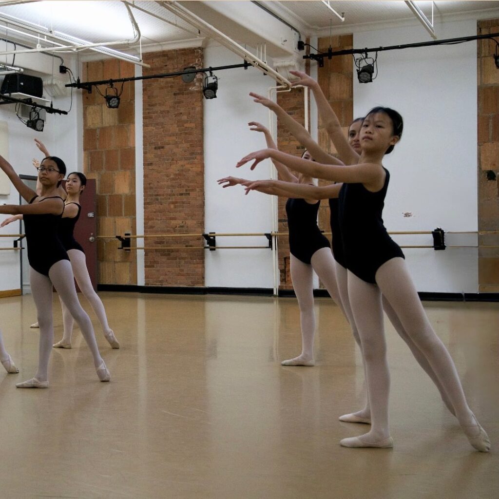A group of students in a ballet studio pose in fourth position with a tendu to the back.