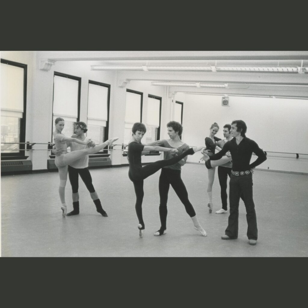 A black and white photo of adult dancers in a dance studio rehearsing with Eliot Feld. Three couples partner each other. The women are on pointe with one leg extended to the front.
