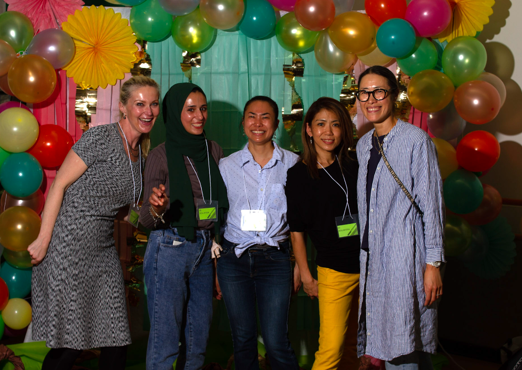 Five parents stand together smiling in front of a colorful backdrop with balloons. They wear visitors passes, participating at a school event.